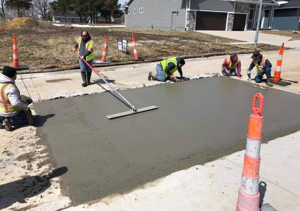 One man is using a tool to flatten a wet concrete slab while four other workers in safety vests are positioned at the sides of the slab.