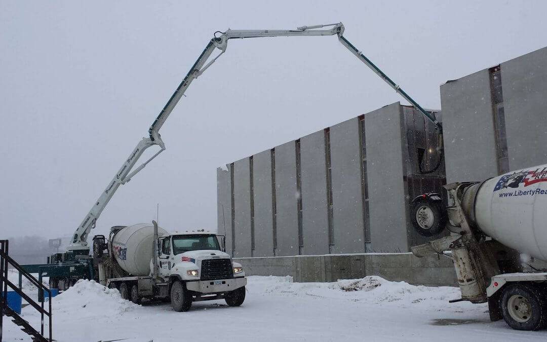 Two white Liberty Ready Mix concrete trucks are parked and pumping concrete in the middle of a snow during winter.